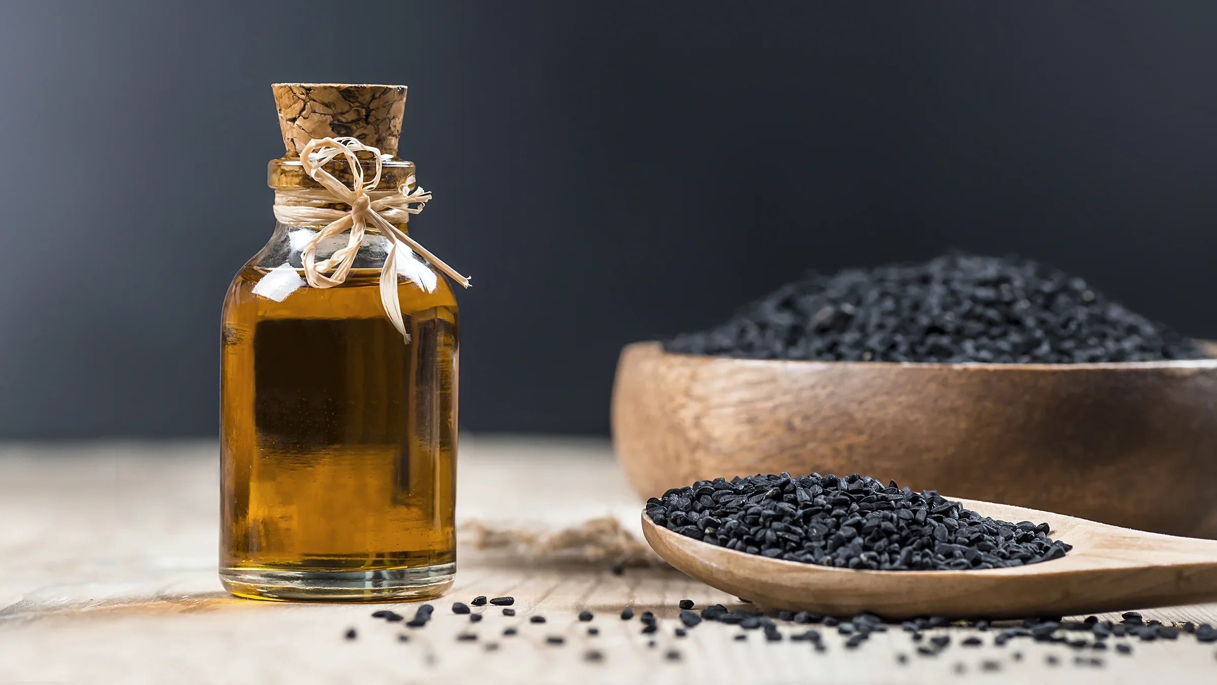 Glass bottle of black seed oil with wooden bowls and spoon of black cumin seeds on table